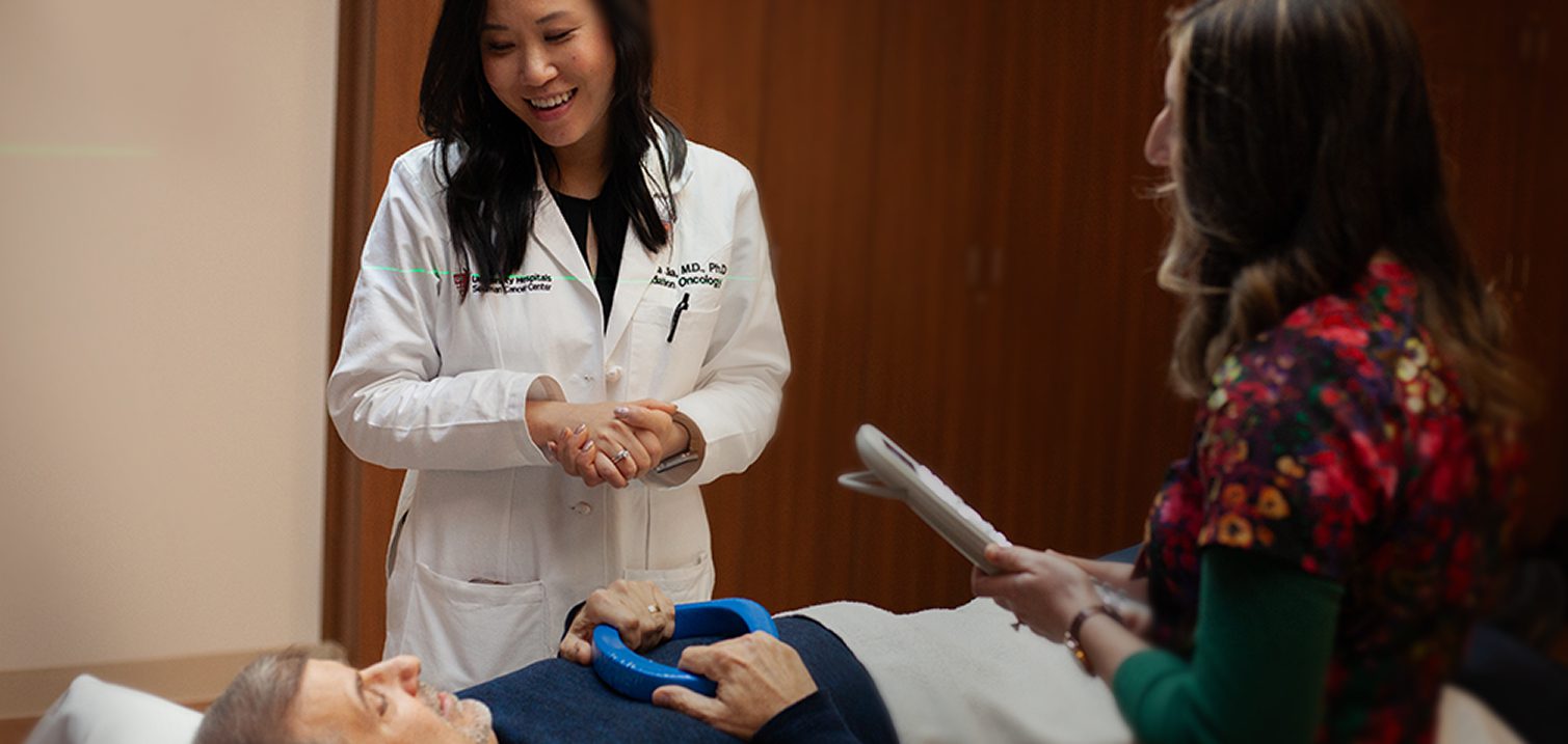 A doctor in a white coat smiling while speaking with a patient in bed and a healthcare professional holding a clipboard.