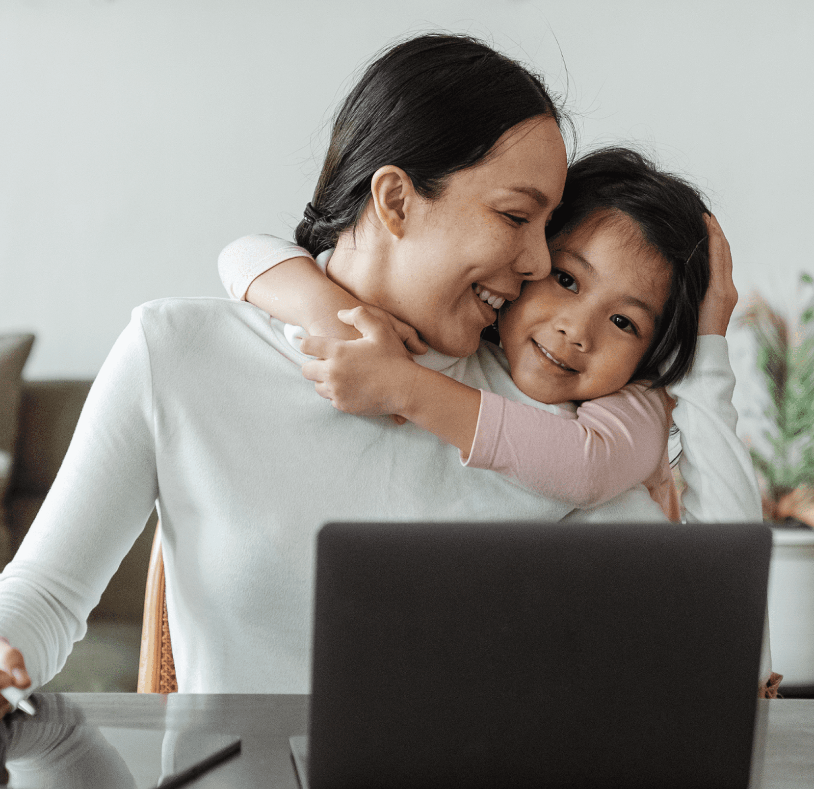 A smiling mother working on a laptop while her young daughter hugs her from behind, creating a loving moment at home.