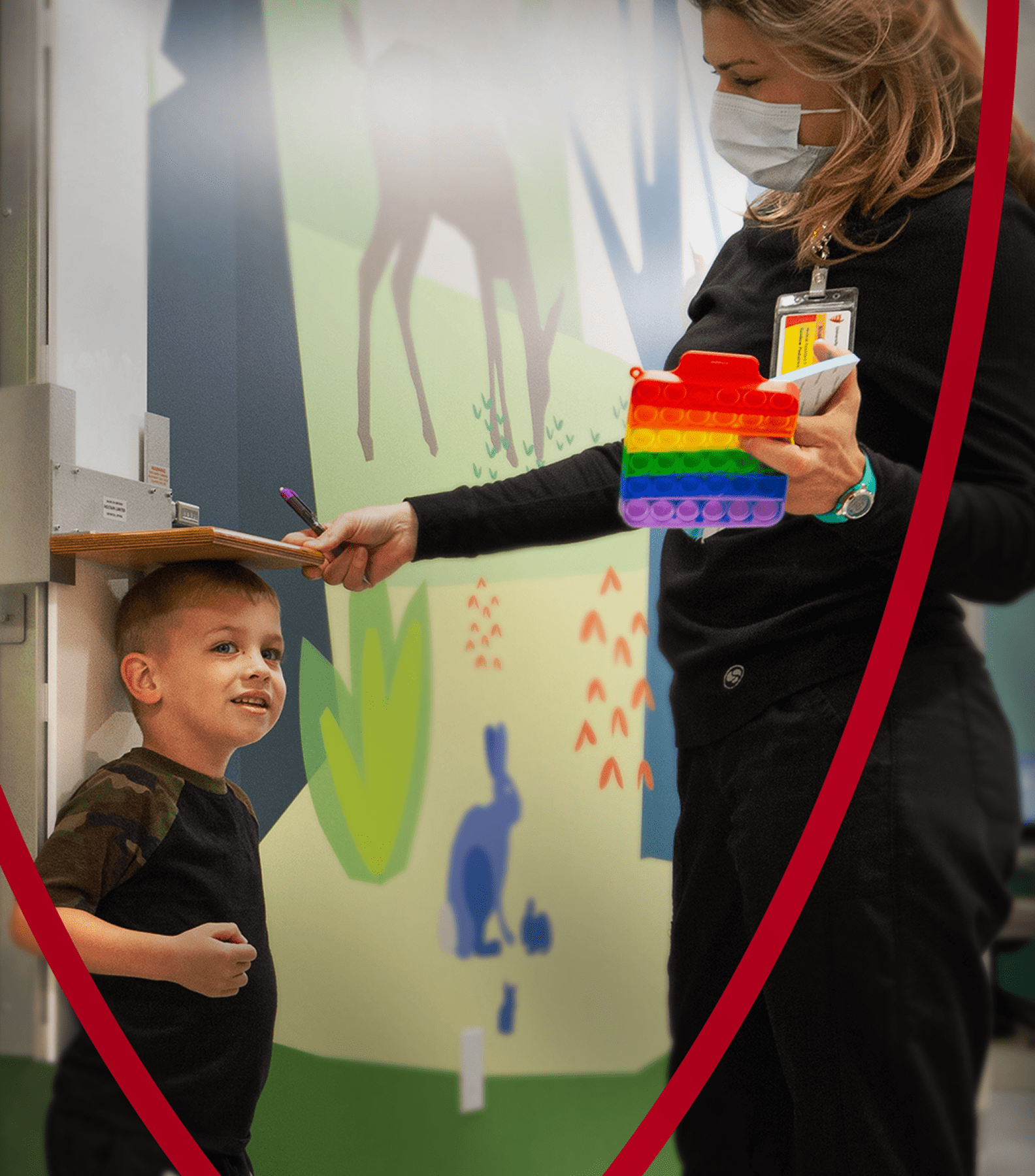A young boy smiles as a masked healthcare professional measures his height in a colorful, child-friendly medical setting.