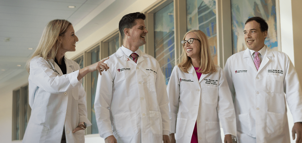 b58d040e04ef11f1414252b9818ef7c7998e611d A group of healthcare professionals in white coats walk together, smiling and engaged in conversation in a hospital corridor.