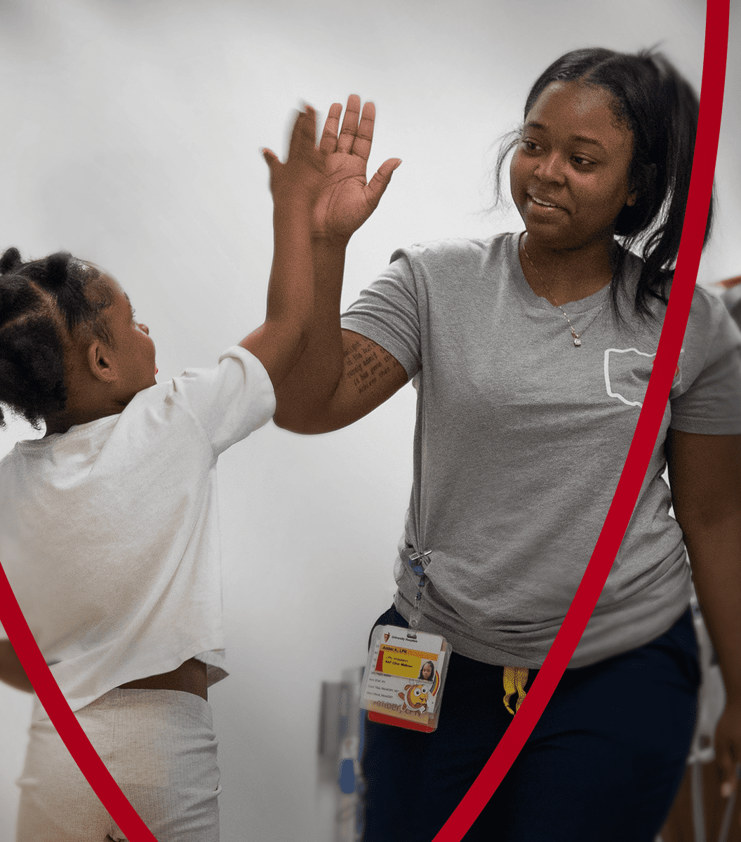 fe5d11e387d643f0db2d793f295537bb09e3fd6a A smiling healthcare worker in a gray shirt high-fives a young child in a hospital hallway.