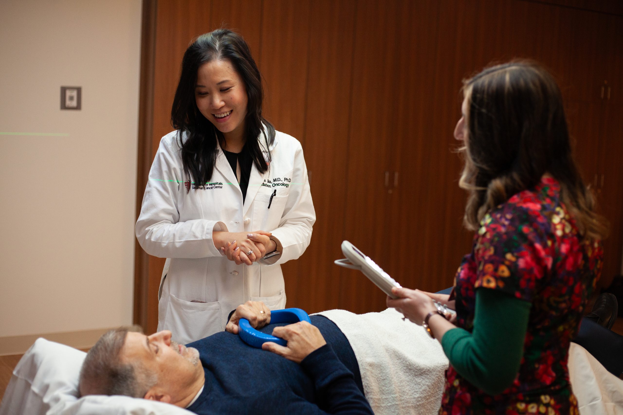 d7b25dd46df944b8cbc994872aef2e52b22cacfb A doctor in a white coat smiling while speaking with a patient in bed and a healthcare professional holding a clipboard.