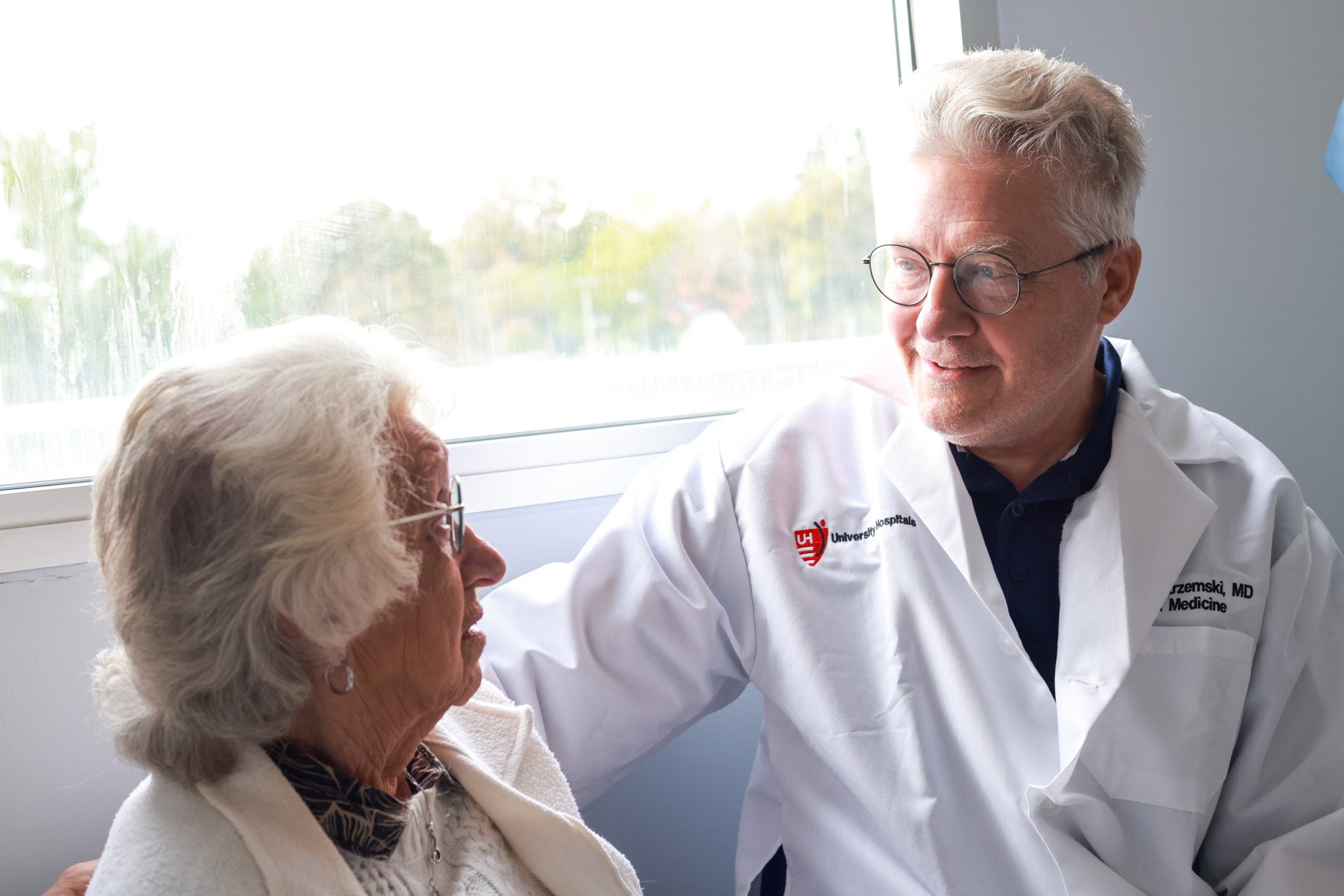 e36084f42fe8170a999605b669aabeaba7d3be54 A doctor in a white coat sits next to an elderly patient, offering compassionate care and conversation.