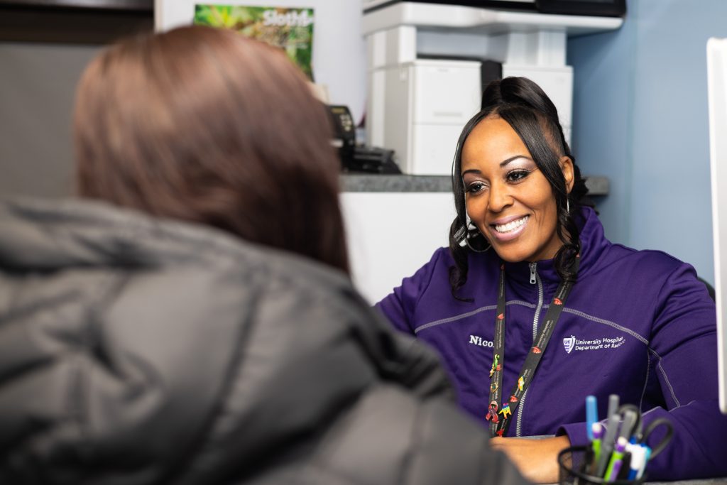 19992e2b1c2089fc18ee64960d436f365f3bbb3d A smiling healthcare professional in a purple University Hospitals jacket engaging warmly with a patient in a clinical setting.