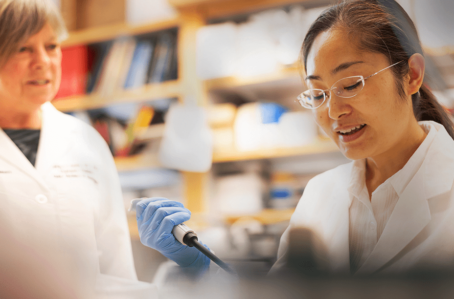 A scientist in a lab coat and gloves using a pipette while a colleague observes, working in a research laboratory setting.