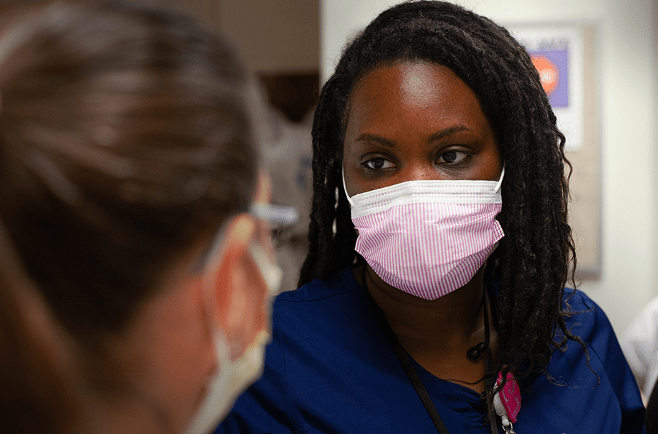 A healthcare professional in navy scrubs wearing a pink face mask attentively talking with a colleague in a clinical setting.