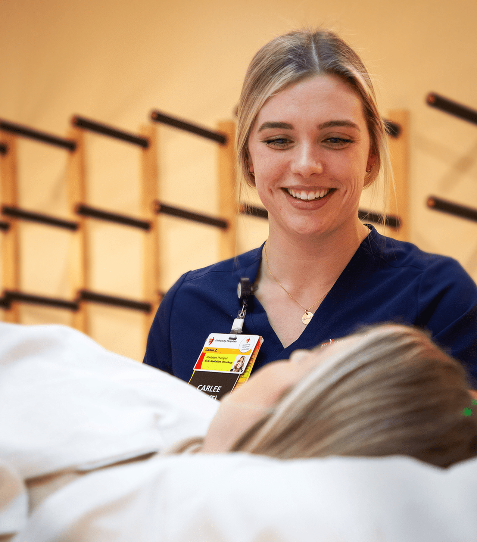 A smiling healthcare professional in navy scrubs interacting with a patient in a medical setting.