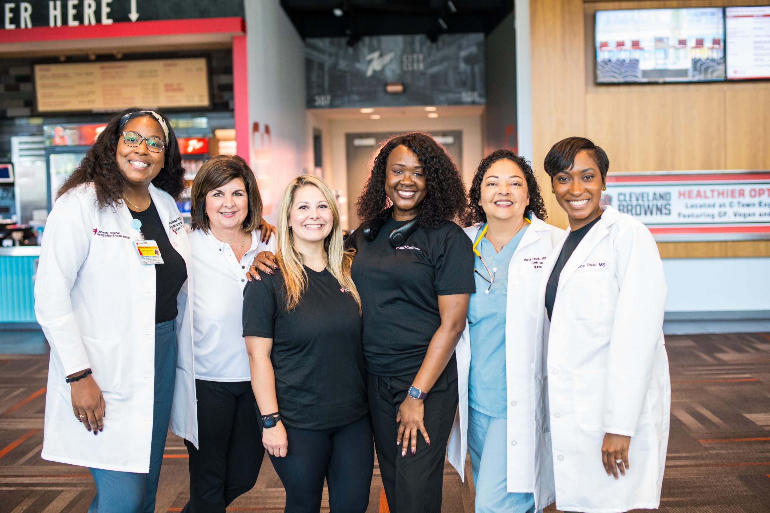  A diverse group of six healthcare professionals—including doctors in white coats and nurses in scrubs—standing side by side in a clinic lobby area, smiling warmly at the camera