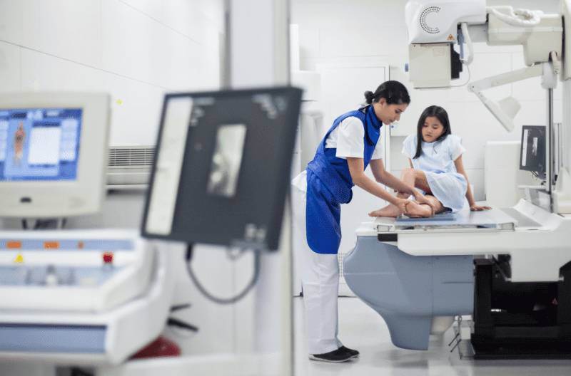 A radiology technician guiding a patient onto an imaging table inside a medical imaging suite equipped with advanced scan equipment